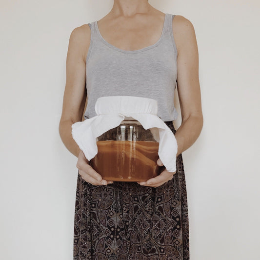 A glass jar of brewing kombucha held in both hands, a cloth tied over the top and the culture resting on the surface of the tea.